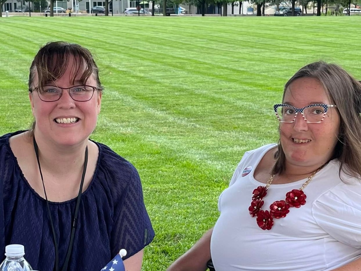 Two women sit at a patriotically decorated information table, in an outdoor, parklike setting.