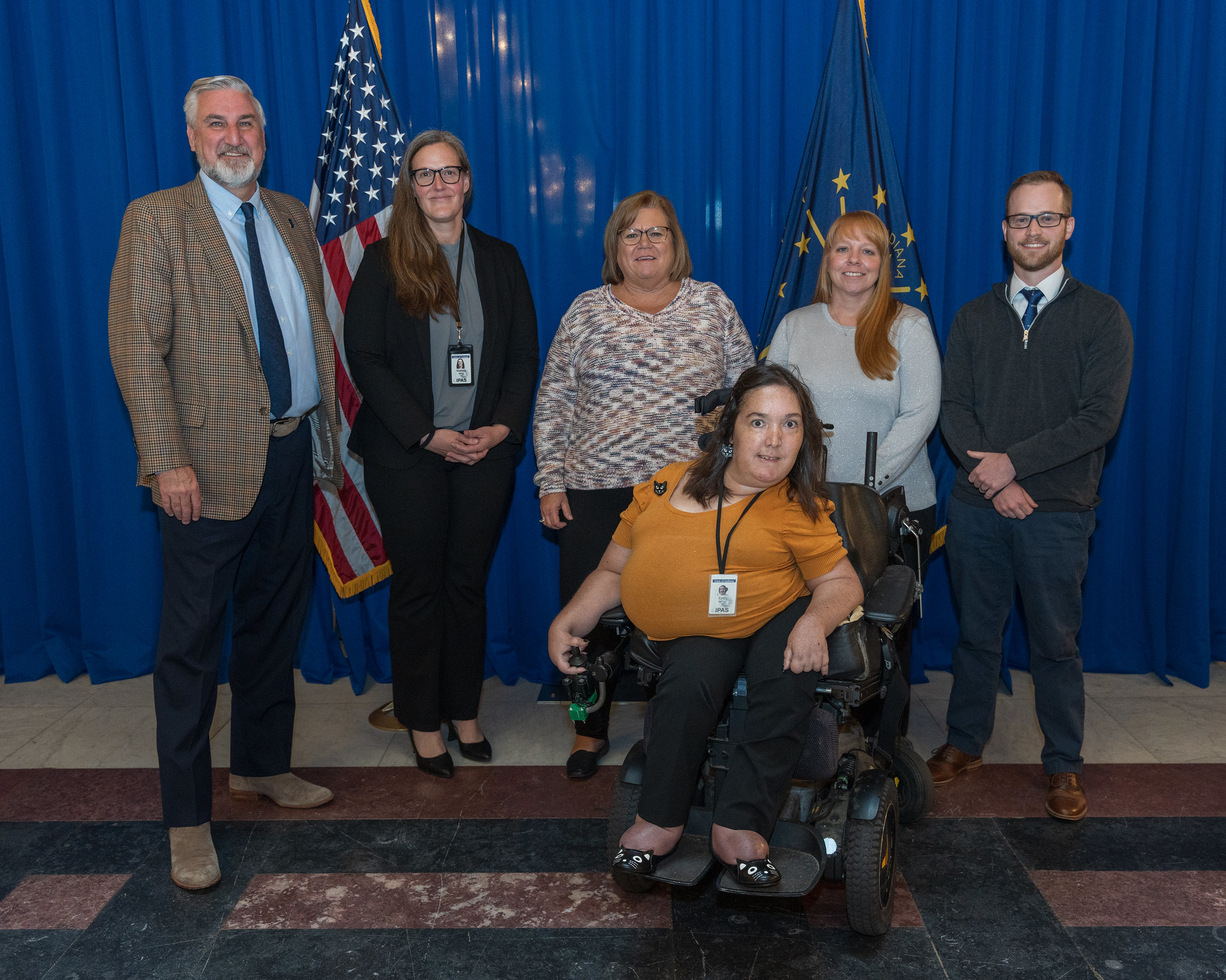 A group of four women and two men pose in front of a seated audience. Behind them are a U.S. flag, the Indiana flag, and a backdrop of blue, draped fabric.