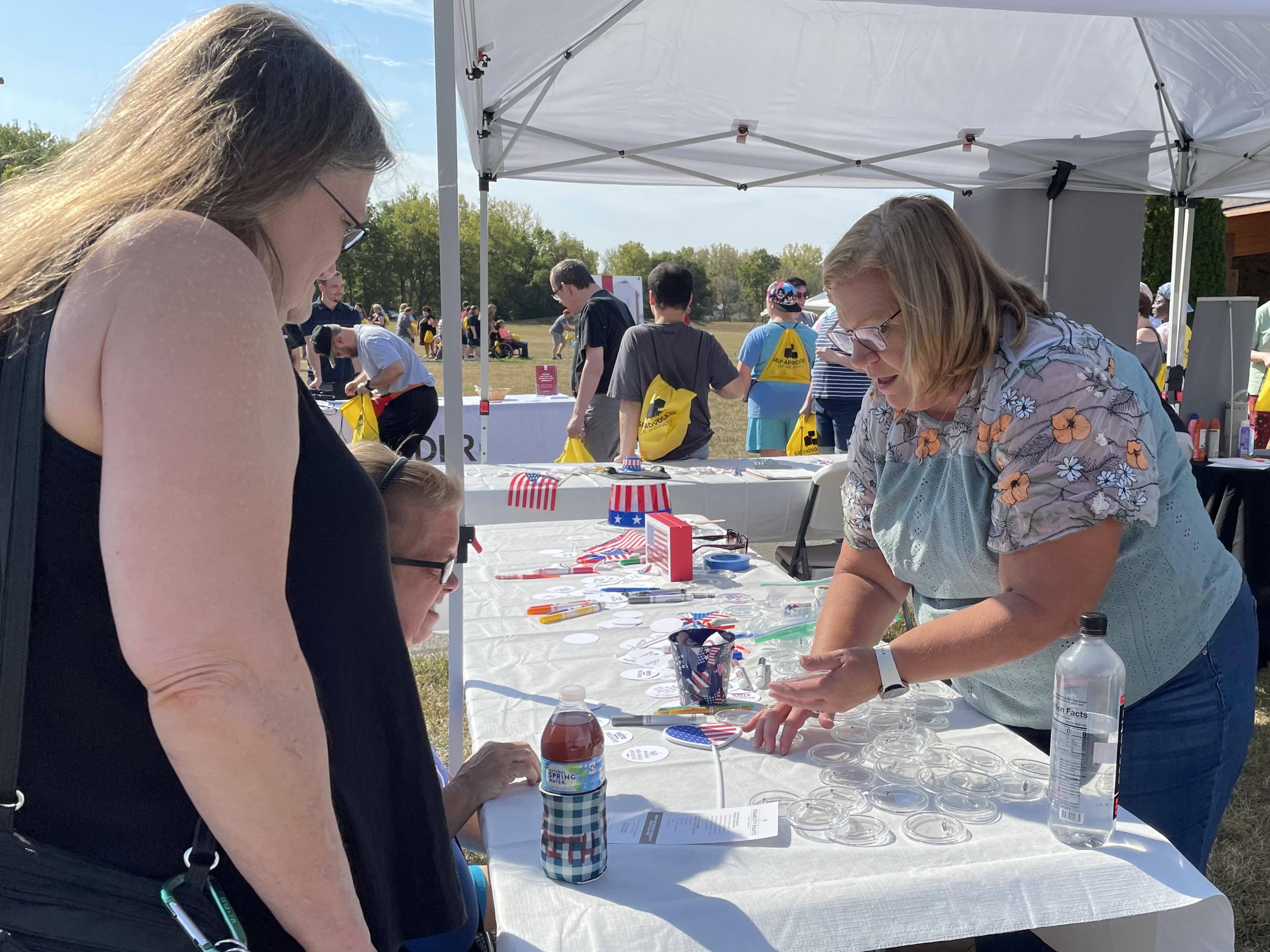 At an outdoor event, three women talk as one creates a craft.