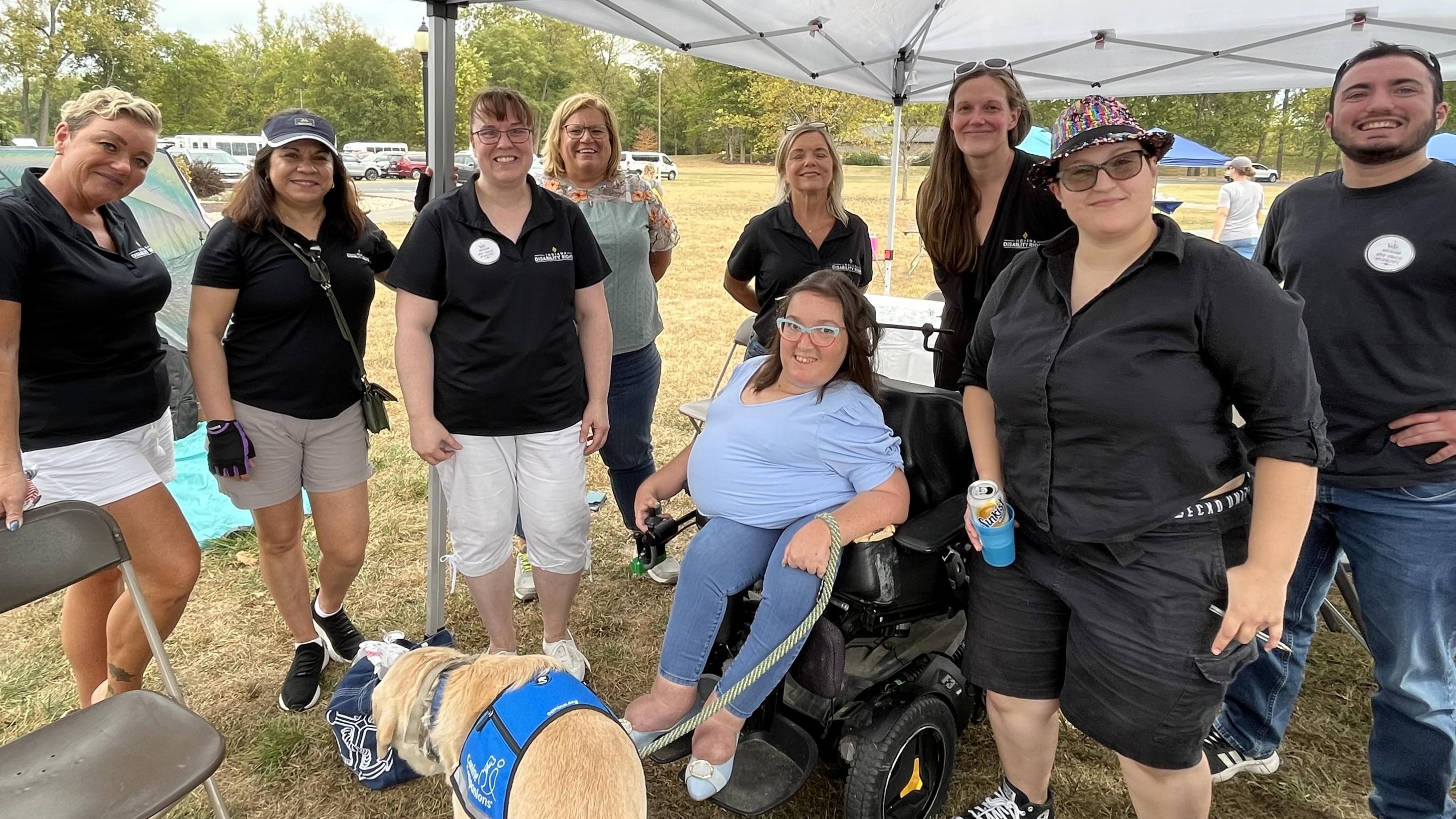 Nine men and women and one service dog, at an outdoor booth, with tables and chairs around and awnings overhead for shade. 