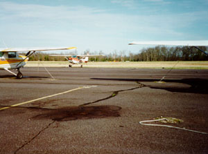 Overview photo of an apron with multiple larger areas of oil staining on the pavement surface.            The photo also shows three smaller airplanes sitting on this apron near the stains.