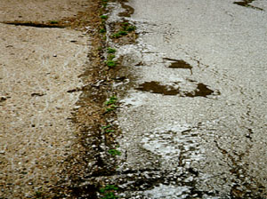 Close-up photo showing a longitudinal joint between a PCC and an asphalt pavement.           The asphalt surface in the photo has a considerable amount of distress that has occurred from shoving.
